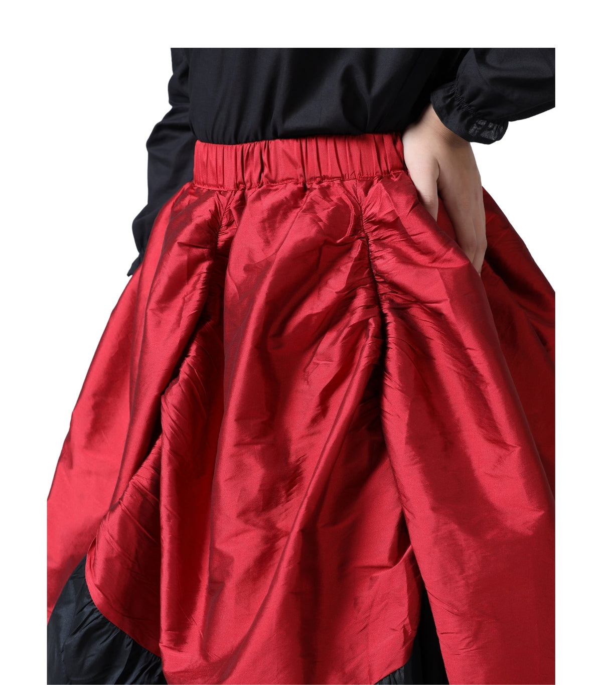 A close-up of a red satin-like skirt with a gathered elastic waistband, worn with a black top. A hand is visible at the side pocket of the skirt against a plain white background.