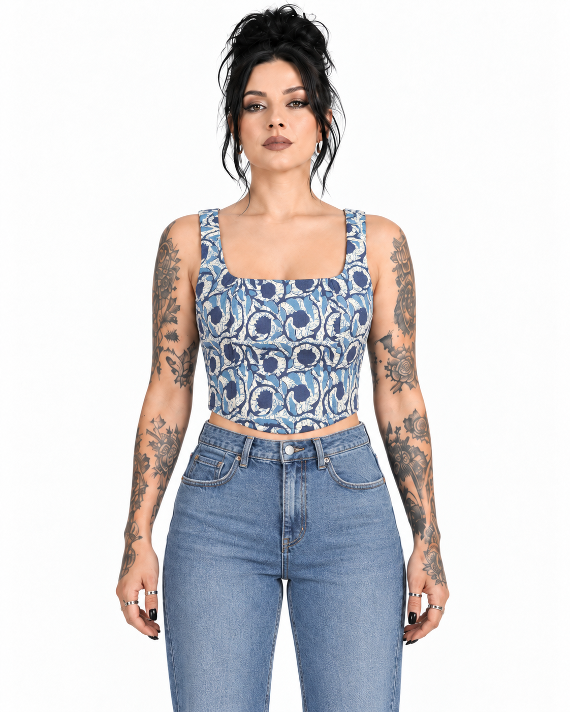 A woman with dark hair and tattoos models a blue and white patterned square-neck crop top with light-wash blue jeans against a plain white background.