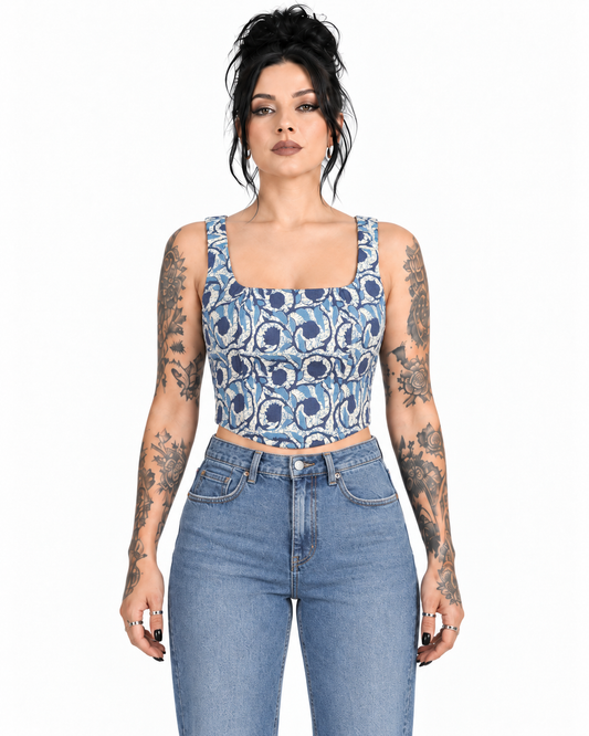 A woman with dark hair and tattoos models a blue and white patterned square-neck crop top with light-wash blue jeans against a plain white background.
